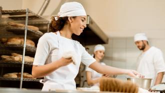 Woman working with dough in commercial bakery with two others standing nearby.